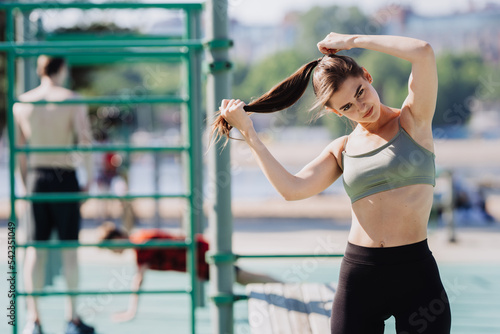 Tired sporty brunette girl at sport court in bra and leggings making ponytail looks at camera. Exhausted spanish girl at her morning training at break. Fitness and health care. European girl trains.