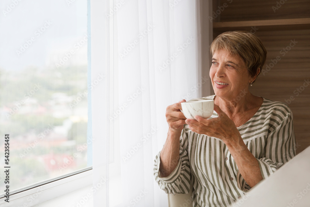 senior woman drinks tea and looks out the window in the kitchen at home. concept without stress. mental health. Slow life. Enjoying the little things