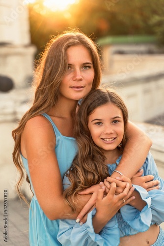 Portrait of mother and daughter in blue dresses with flowing long hair against the backdrop of sunset. The woman hugs and presses the girl to her. They are looking at the camera.