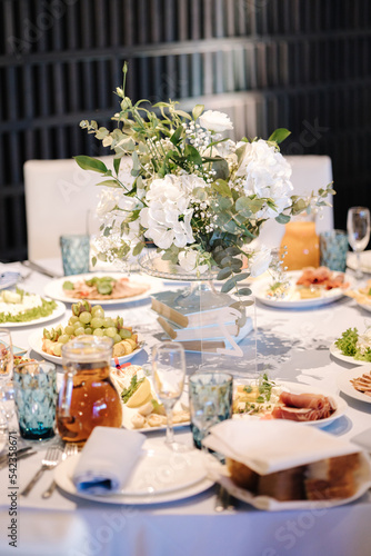 Big round table decorated by white and blue flowers in restaurant. Different dishes on table. Cristening of little boy. Luxury decor