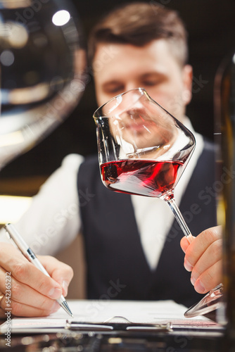 Wine steward holds wineglass with red drink, writes review.