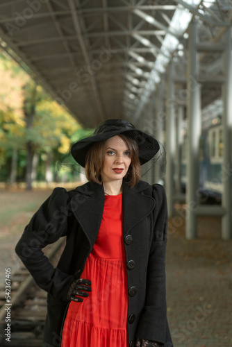 Portrait of middle-aged aristocratic woman in red dress, black coat and hat standing on platform of station waiting for train © somemeans
