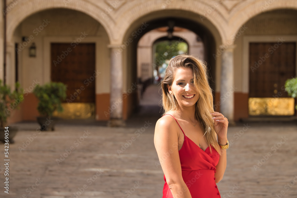 Portrait of young, attractive, blonde woman in an elegant red party dress touching her hair while smiling around the city. Concept beauty, fashion, elegance, luxury. Portrait of young, attractive, blonde woman in an elegant red party dress touching her hair while smiling around the city. Concept beauty, fashion, elegance, luxury.