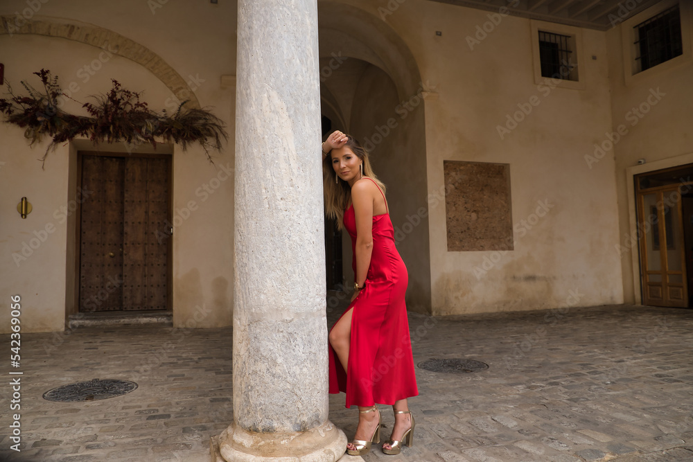 Young, attractive, blonde woman in an elegant red party dress leaning on a marble column smiling happily. Concept beauty, fashion, elegance, luxury.
