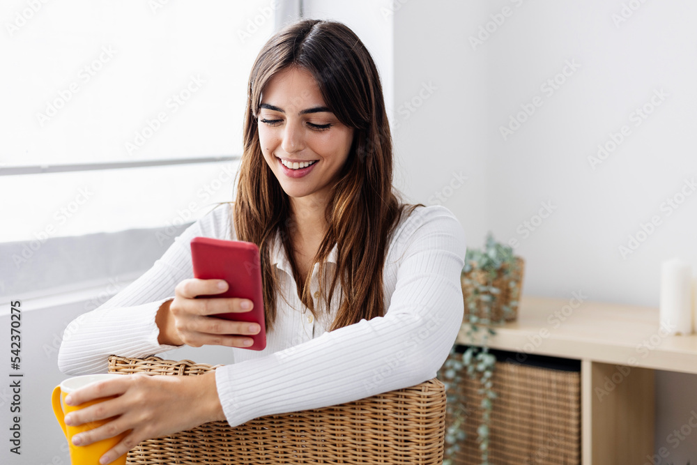 Smiling woman holding coffee cup using smart phone on chair at home Smiling woman holding coffee cup using smart phone on chair at home