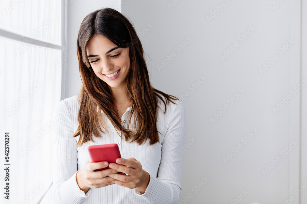 Smiling woman using smart phone by window at home Smiling woman using smart phone by window at home