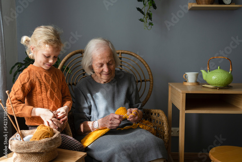 Smiling girl holding ball of wool standing by grandmother knitting on chair at home photo