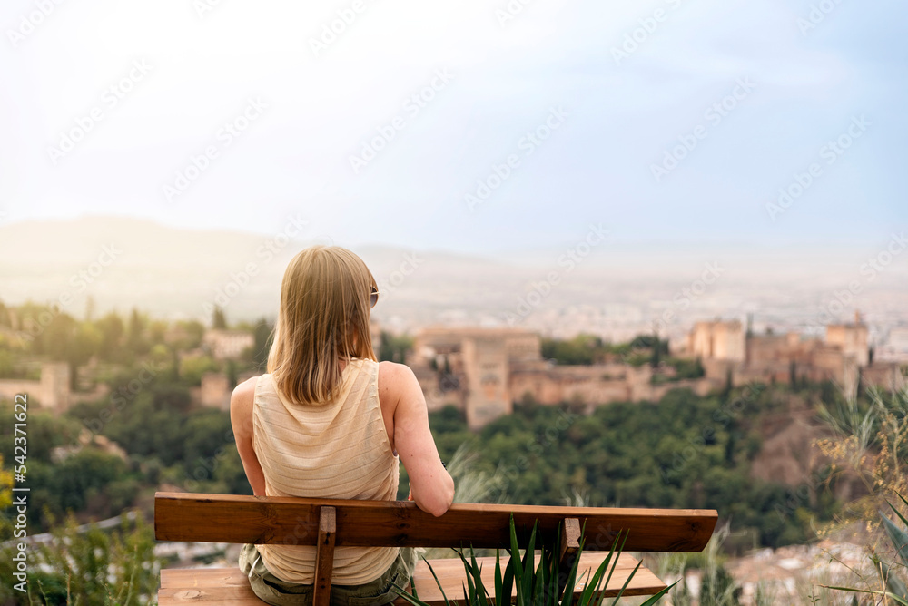 A young female traveler sitting on a bench contemplates the views of the Alhambra palace in the city of Granada, Spain