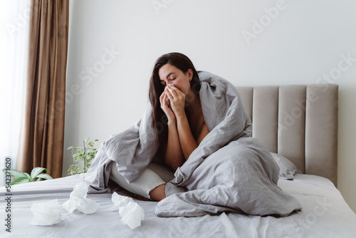 Ill woman blowing nose in tissue on bed at home photo