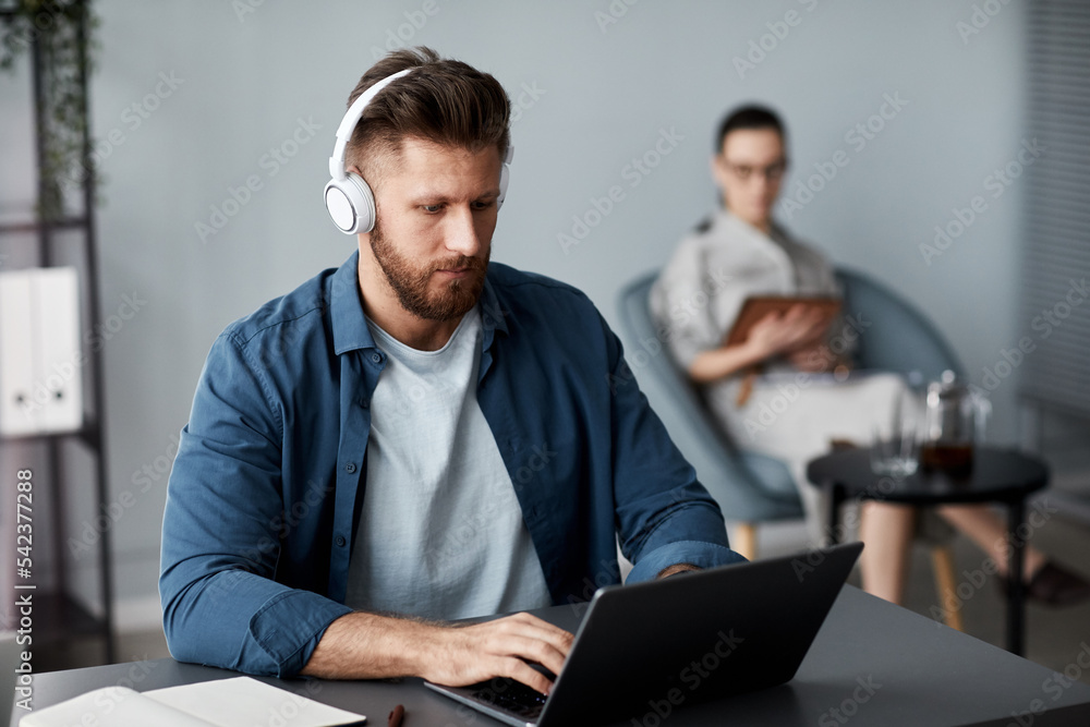 Young serious man in headphones and casualwear sitting in front of laptop and carrying out online assignment against woman