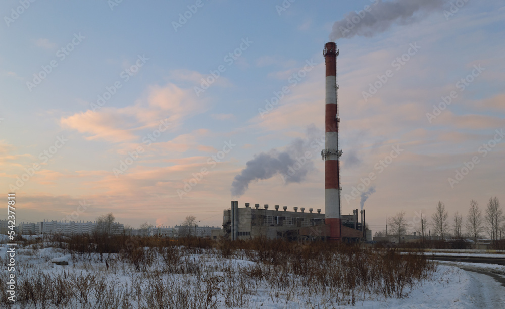 Boiler room with a tall striped smoking chimney in the middle of a winter evening landscape