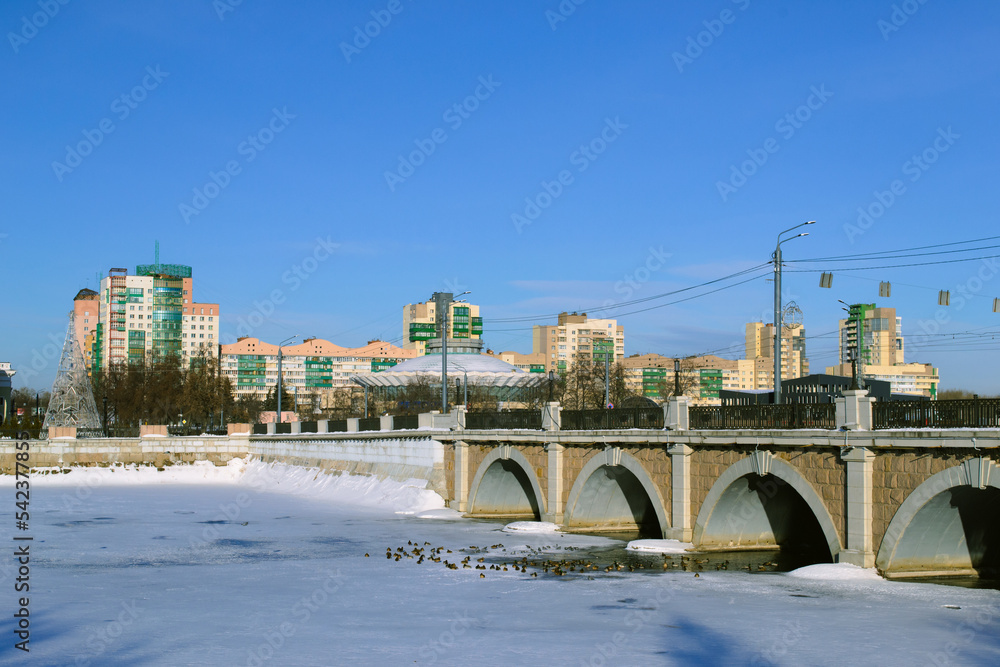 Naklejka premium Close-up of the historic arch bridge and the frozen river below it against the backdrop of urban residential buildings