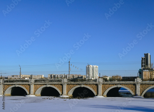 Close-up of the historic arch bridge and the frozen river below it against the backdrop of urban residential buildings © Maksim