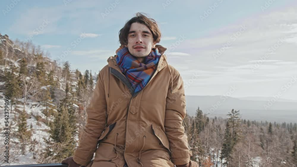 Handsome young Caucasian man sitting relaxed on protection fence at viewing platform on mountain top looking away then looking at camera Handsome young Caucasian man sitting relaxed on protection fence at viewing platform on mountain top looking away then looking at camera