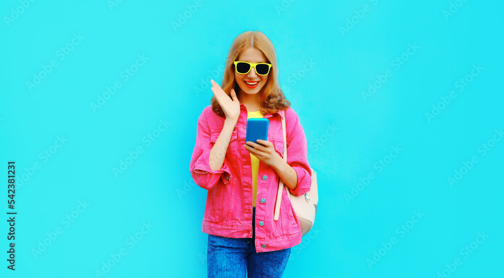 Portrait of stylish modern happy smiling young woman with smartphone wearing pink jacket, backpack on blue background