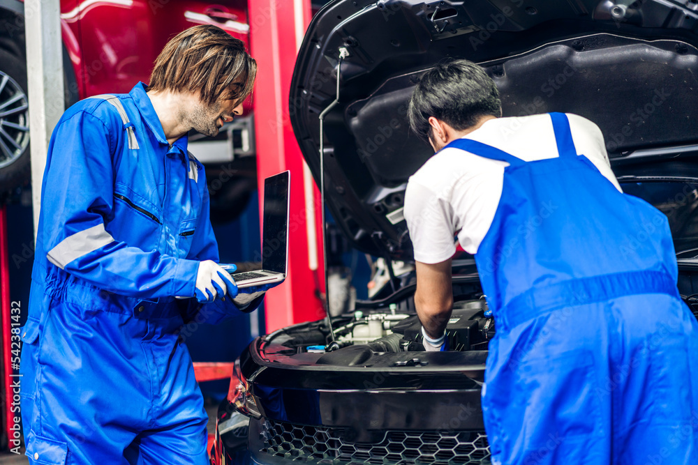 Professional car technician mechanic team in uniform use laptop work fixing vehicle car engine and maintenance repairing checking under car hood in auto service. Automobile service garage Professional car technician mechanic team in uniform use laptop work fixing vehicle car engine and maintenance repairing checking under car hood in auto service. Automobile service garage