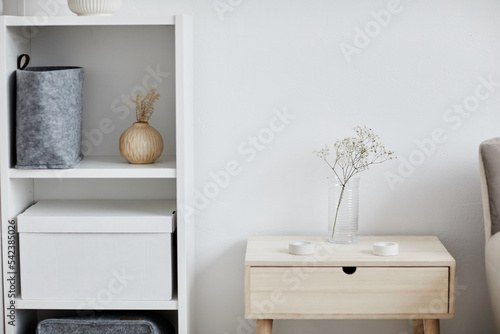 Part of cozy bedroom with dried flowers in transparent vase standing on nightstand and shelf with large white box by white wall