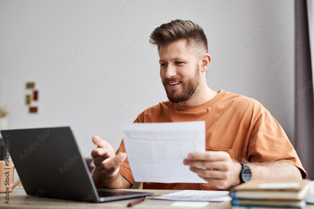 Happy young tutor or student with document sitting in front of laptop at home and looking at screen during video communication Happy young tutor or student with document sitting in front of laptop at home and looking at screen during video communication