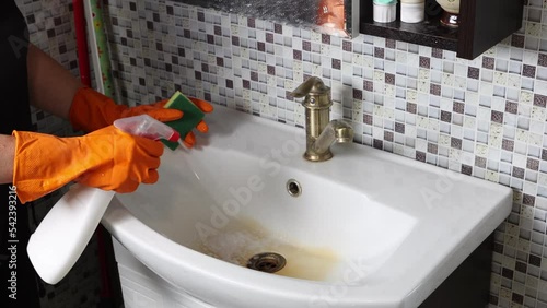 Cleaning lady cleans ceramic sink in bathroom by spraying detergent and wiping with sponge.