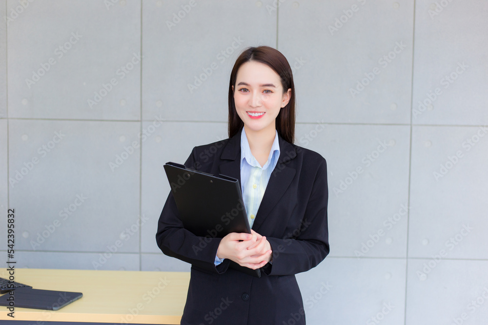 Young Asian professional working woman in a black suit holds clipboard in her hands and confident smiles in office room. Young Asian professional working woman in a black suit holds clipboard in her hands and confident smiles in office room.