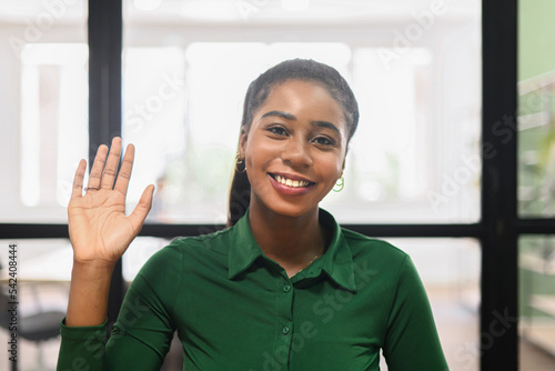 Friendly adorable African American female office worker businesswoman freelancer with Afro hairstyle joyfully waving, smiling and saying hi, making hello sign, greeting and welcoming a newcomer photo