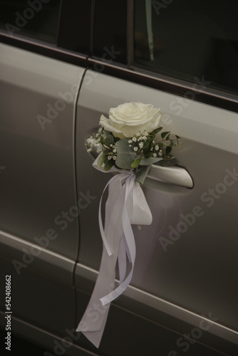 A close-up of a wedding car decorated with a white rose, greenery, and a flowing ribbon.