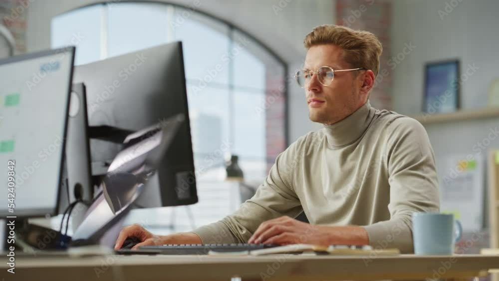 Portrait of White Creative Young Man With Glasses Working on a Computer During Day Time in a Spacious Modern Office. Male Graphic Designer Smiling While Developing a New Visual Concept. Dolly Shot. Portrait of White Creative Young Man With Glasses Working on a Computer During Day Time in a Spacious Modern Office. Male Graphic Designer Smiling While Developing a New Visual Concept. Dolly Shot.