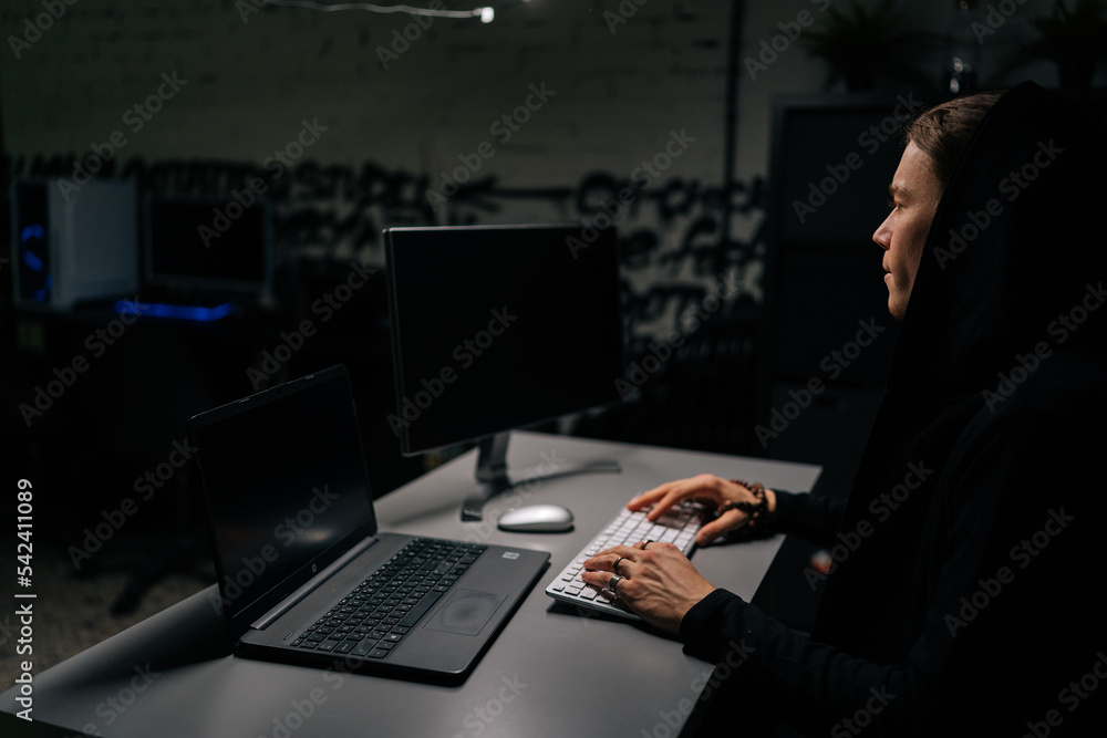 Side view of dangerous hacker man wearing hooded typing on wireless keyboard breaking password, looking at laptop and desktop screen. Concept of data thief, internet attack, darknet and cyber security