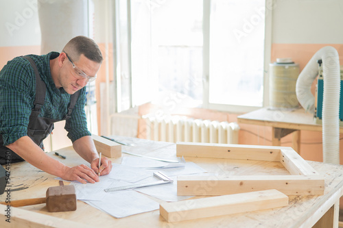 A portrait of a carpenter in goggles and work overalls draws a blueprint for a workshop.