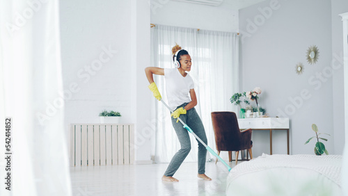 Cheerful African American girl in gloves is mopping floor, listening to music with wireless headphones and dancing enjoying rhythm cleaning her nice modern apartment.