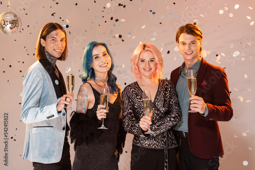 joyful nonbinary people with champagne glasses looking at camera while celebrating christmas on beige background