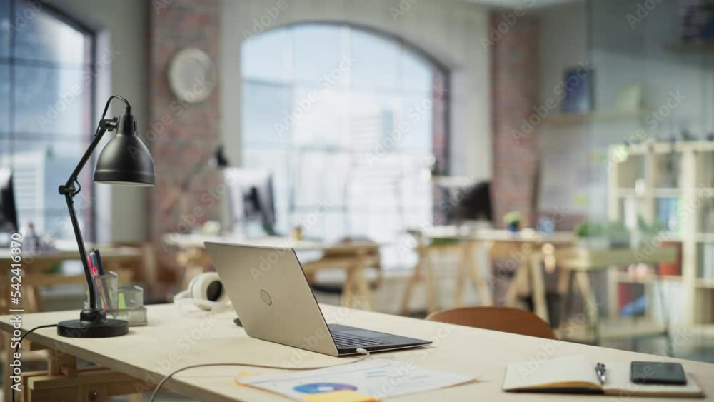 Charging Laptop on a Desk in a Bright Spacious Empty Workplace. Office Supplies Are on the Table. Big Windows with City Skyscrapers View in the Background. Slow Dolly arc Shot. Charging Laptop on a Desk in a Bright Spacious Empty Workplace. Office Supplies Are on the Table. Big Windows with City Skyscrapers View in the Background. Slow Dolly arc Shot.