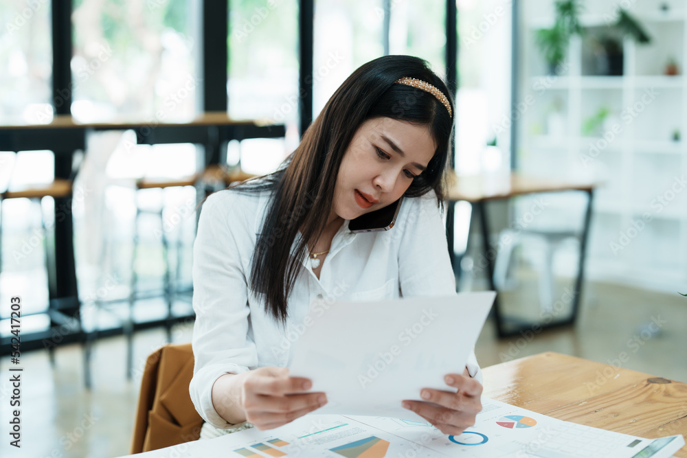 A portrait of a beautiful Asian female employee showing a stressed face while using the phone and financial documents on her desk A portrait of a beautiful Asian female employee showing a stressed face while using the phone and financial documents on her desk