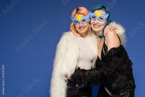 joyful queer people in party masks and faux fur jackets embracing isolated on blue