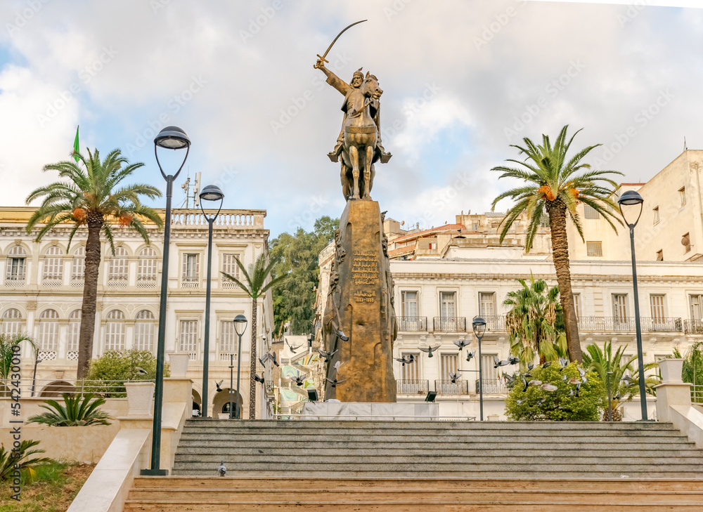 El Amir Abdelkader statue with flying pigeons and palm trees in Emir abdelkader place, Larbi Ben M'hidi and Colonel Haouas road. Algiers city town hall building. Stairs, posts, trees and cloudy sky.