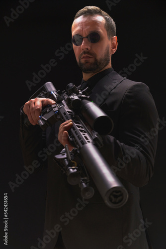 Secret service man in black suit, bodyguard or special forces agent man with gun isolated on black background. Man in black suit holding assault rifle. Spy, police man in black suit and sunglasses