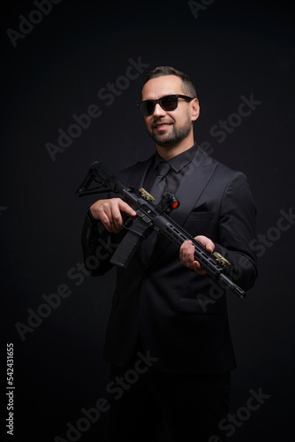 Self confident man with rifle in black suit and sunglasses. Man holding assault rifle. Secret service man in black suit, bodyguard or special forces agent man with gun isolated on black background