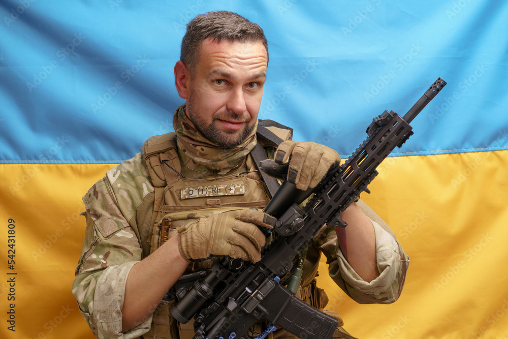 Soldier of Ukrainian Army handsome man holding rifle in a camouflage ammunition posing on Ukrainian flag background. Brave Ukrainian army man with weapon. The flag of Ukraine on background Soldier of Ukrainian Army handsome man holding rifle in a camouflage ammunition posing on Ukrainian flag background. Brave Ukrainian army man with weapon. The flag of Ukraine on background