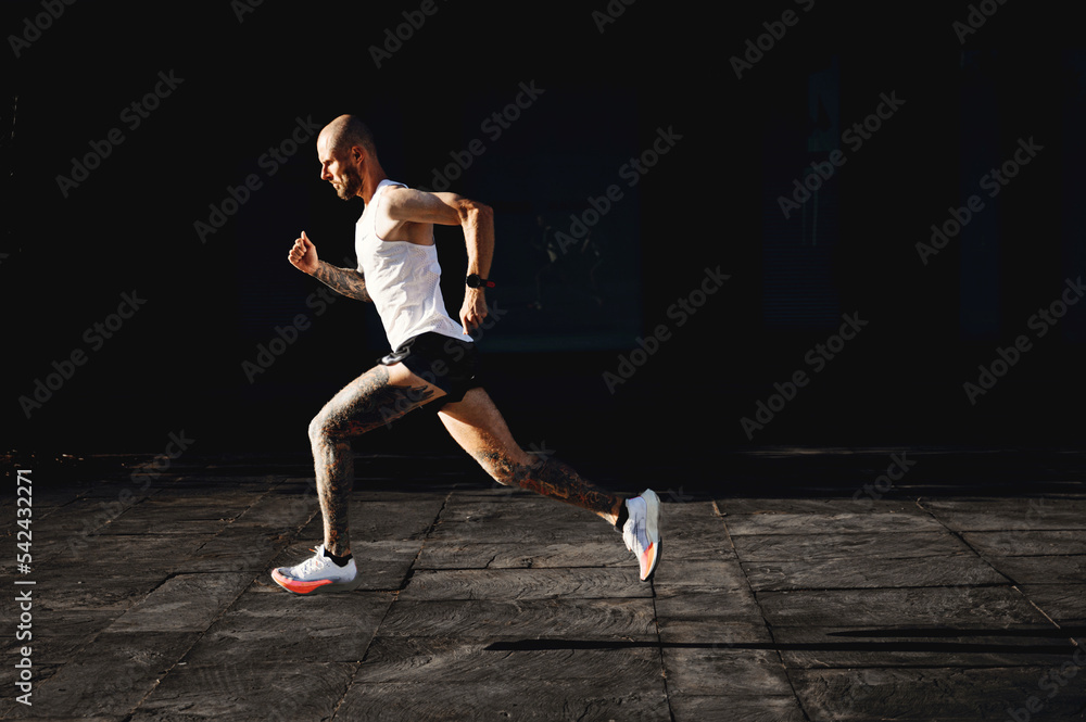 Bald young athletic tattooed man running fast outdoor, sportsman preparing to marathon, doing cardio exercises on the open air, side view