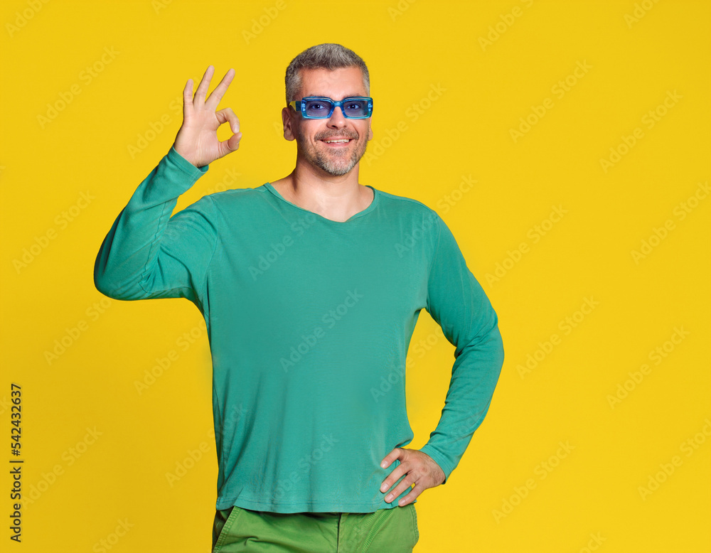 Gesturing OK handsome middle aged man posing on yellow background wearing casual. Grey haired handsome man in blue sunglasses. Fit mature man hand gesture Gesturing OK handsome middle aged man posing on yellow background wearing casual. Grey haired handsome man in blue sunglasses. Fit mature man hand gesture