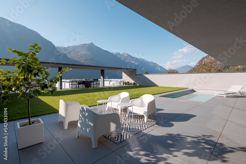Modern house with garden. In the foreground is a small living room with armchairs and small tables  at the back is the pool with a deck chair © alexandre zveiger