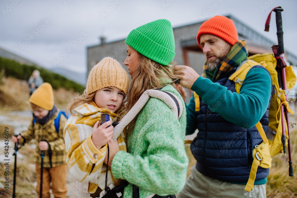 Obraz premium Man helping his wife with their little tired daughter, giving her into child carrier, during autumn hike in mountains.