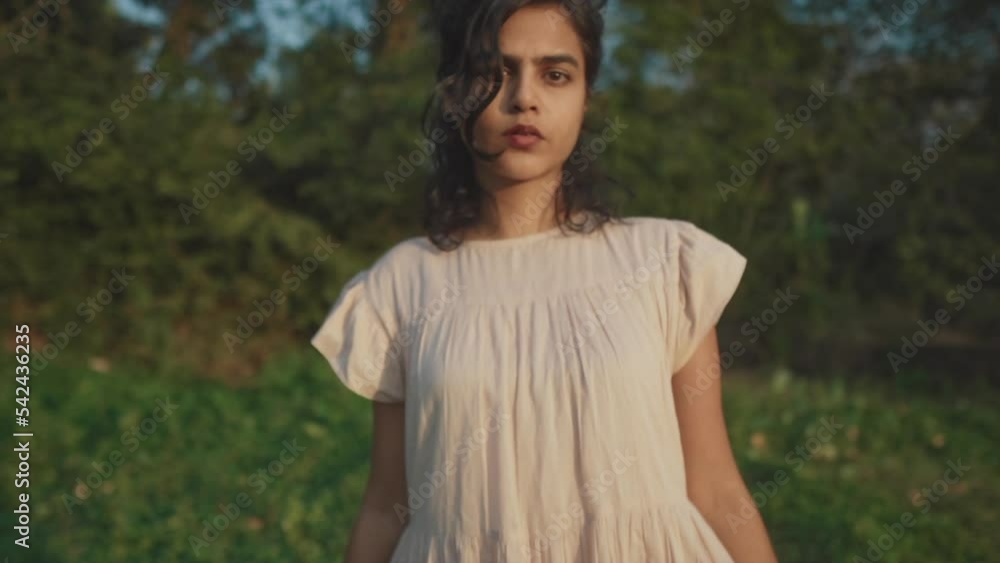 Slow motion backward dolly shot of a young charming indian woman in a dress looking at the camera with a serious look while she is in nature Slow motion backward dolly shot of a young charming indian woman in a dress looking at the camera with a serious look while she is in nature