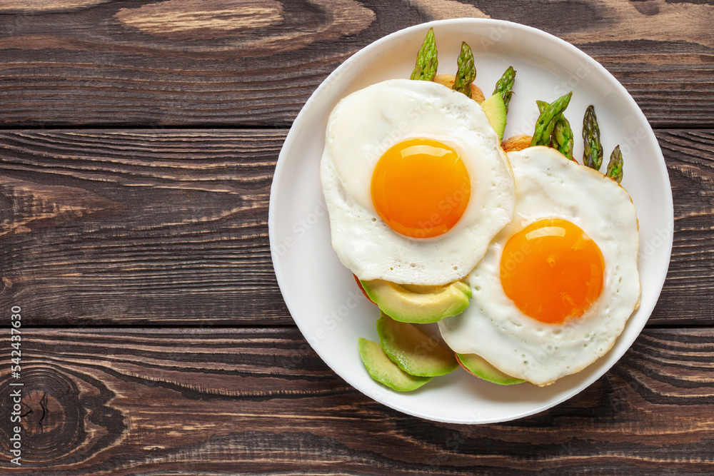 Tasty breakfast. Fried eggs with green asparagus and avocado on bread toasts on the plate on wooden table. Top view Tasty breakfast. Fried eggs with green asparagus and avocado on bread toasts on the plate on wooden table. Top view