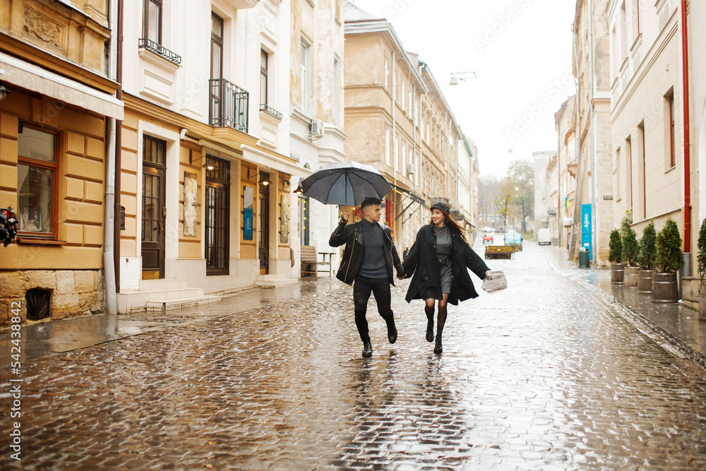 Happy couple running down the street in rainy weather. The guy holds an umbrella in his hand. 