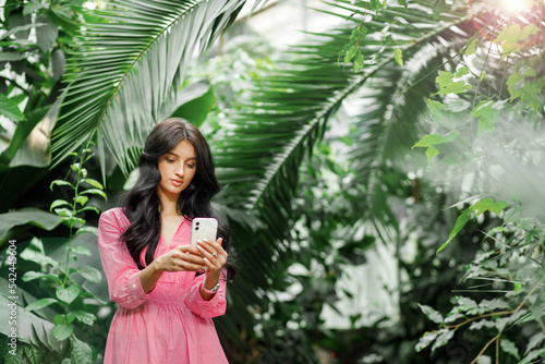 Portrait of young beautiful woman with long wavy brunette hair with her phone in tropical botanic garden with greenery on the background.