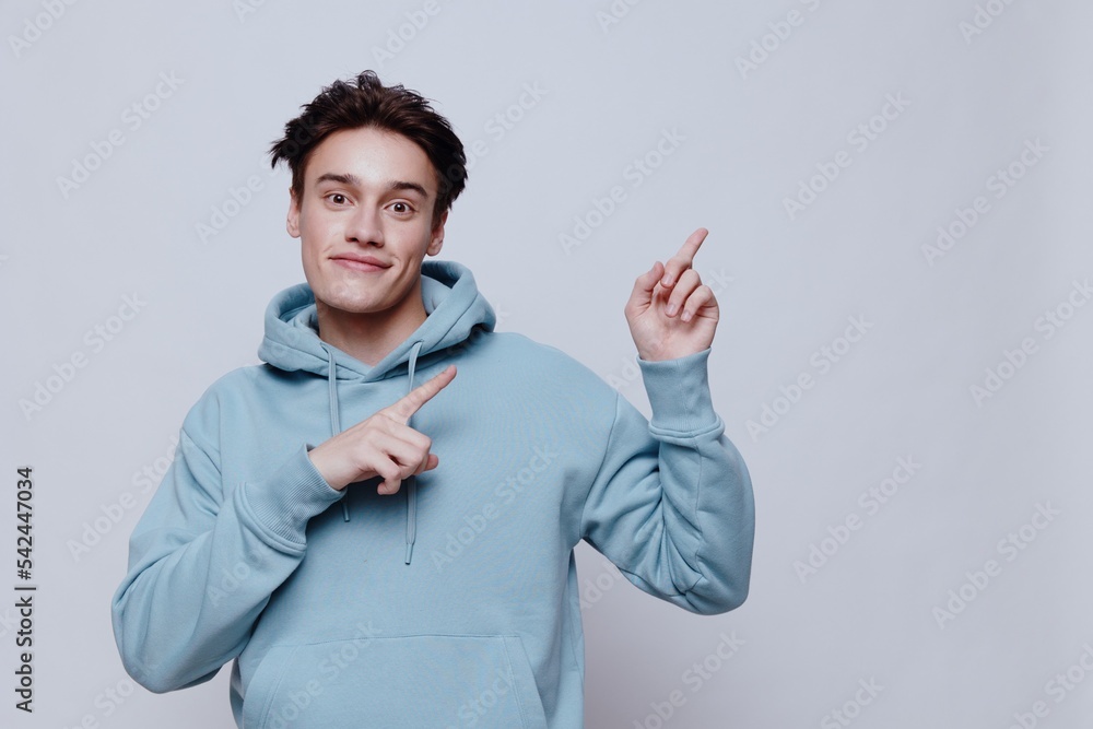 horizontal portrait of a pleasantly smiling young man in a light hoodie pointing his fingers towards an empty space on the background for inserting an advertising layout horizontal portrait of a pleasantly smiling young man in a light hoodie pointing his fingers towards an empty space on the background for inserting an advertising layout