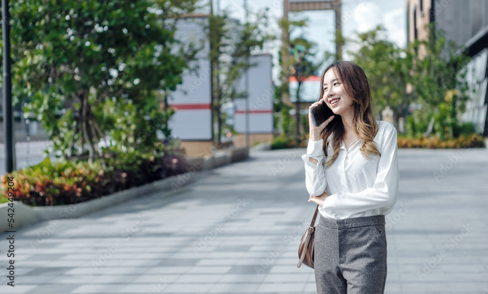Smiling Asian curly business woman wearing trendy walks down the central city street and uses her phone. Pretty summer woman in white shirt walks down the street looking at her mobile phone Smiling Asian curly business woman wearing trendy walks down the central city street and uses her phone. Pretty summer woman in white shirt walks down the street looking at her mobile phone