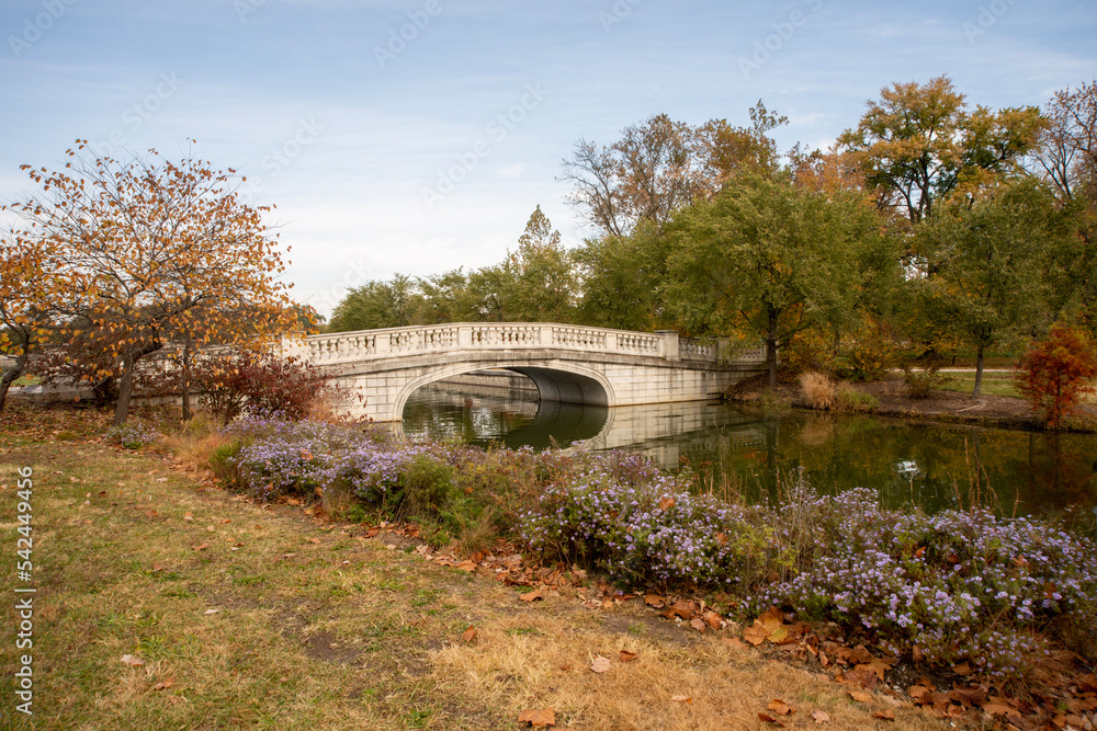 White stone pedestrian bridge with a lake below. Tiny purple flowers line the shore and trees of red, green, orange and yellow can be seen in the autumn. White stone pedestrian bridge with a lake below. Tiny purple flowers line the shore and trees of red, green, orange and yellow can be seen in the autumn.
