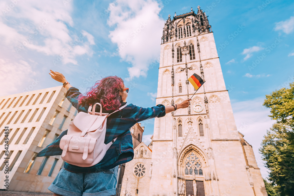 A young happy tourist or student girl with a German flag at the old town or Altstadt in Munster with church belfry in background. Studying language abroad and traveling concept